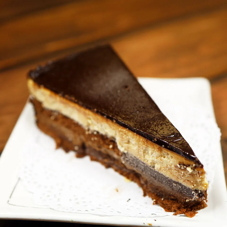 A slice of layered chocolate mousse cake on a white plate with a paper doily, showing distinct layers of dark chocolate glaze, light mousse, and dark sponge cake base at Unique Cafe, a Japanese restaurant in Ontario