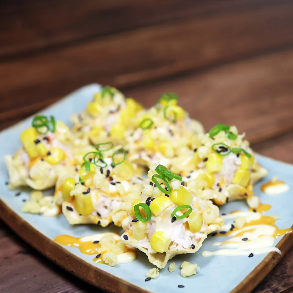 Six bite-sized crispy wonton cups filled with creamy crab salad, topped with sweet corn kernels, chopped green onions, and black sesame seeds, drizzled with spicy mayo on a blue plate at Unique Cafe, a Japanese restaurant in Ontario