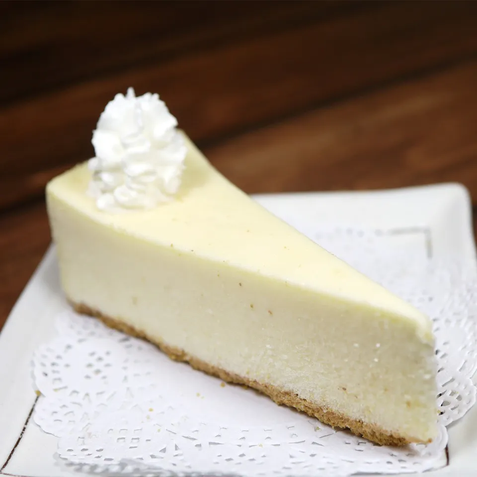 A slice of smooth, pale yellow cream cheesecake with a golden graham cracker crust, topped with a dollop of whipped cream and served on a white plate with a paper doily at Unique Cafe, a Japanese restaurant in Ontario