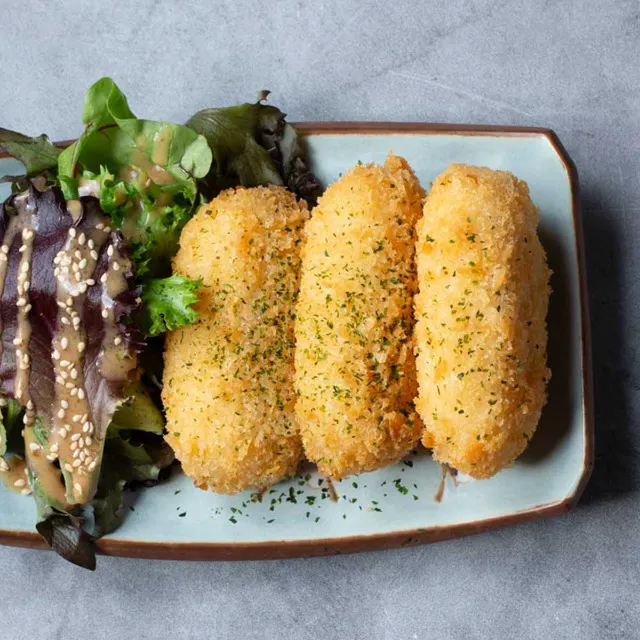 Three oblong, golden-brown breaded cream crab croquettes dusted with parsley herbs, arranged next to a side salad with sesame dressing on a light blue rectangular plate at Unique Cafe, a Japanese restaurant in Ontario