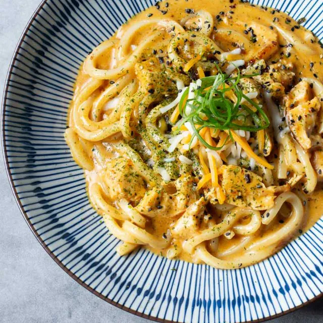 A top-down view of thick udon noodles tossed in a rich, orange-colored cream curry sauce, topped with shredded cheddar cheese, sliced green onions, and black sesame seeds in a blue-striped bowl at Unique Cafe, a Japanese restaurant in Ontario