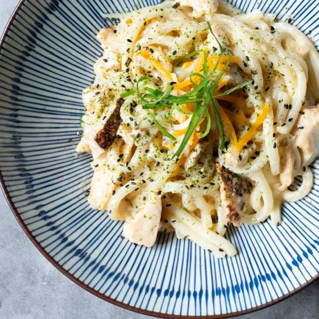 A blue-striped bowl containing thick udon noodles in a white cream sauce mixed with pieces of grilled chicken, sprinkled with shredded cheese, green onions, and black sesame seeds at Unique Cafe, a Japanese restaurant in Ontario