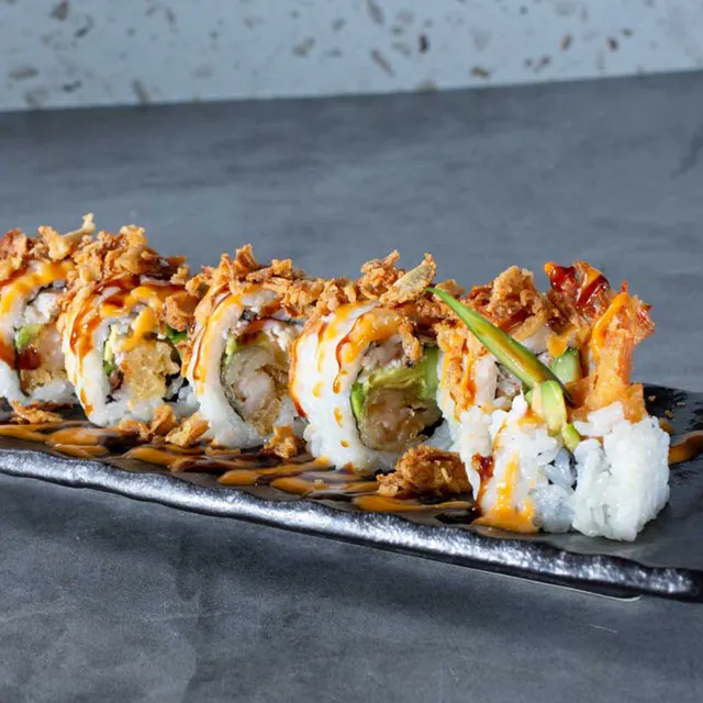 A row of sushi dragon rolls filled with shrimp tempura and avocado, topped with crispy fried onions, and drizzled with eel sauce and spicy mayo on a long black plate at Unique Cafe, a Japanese restaurant in Ontario
